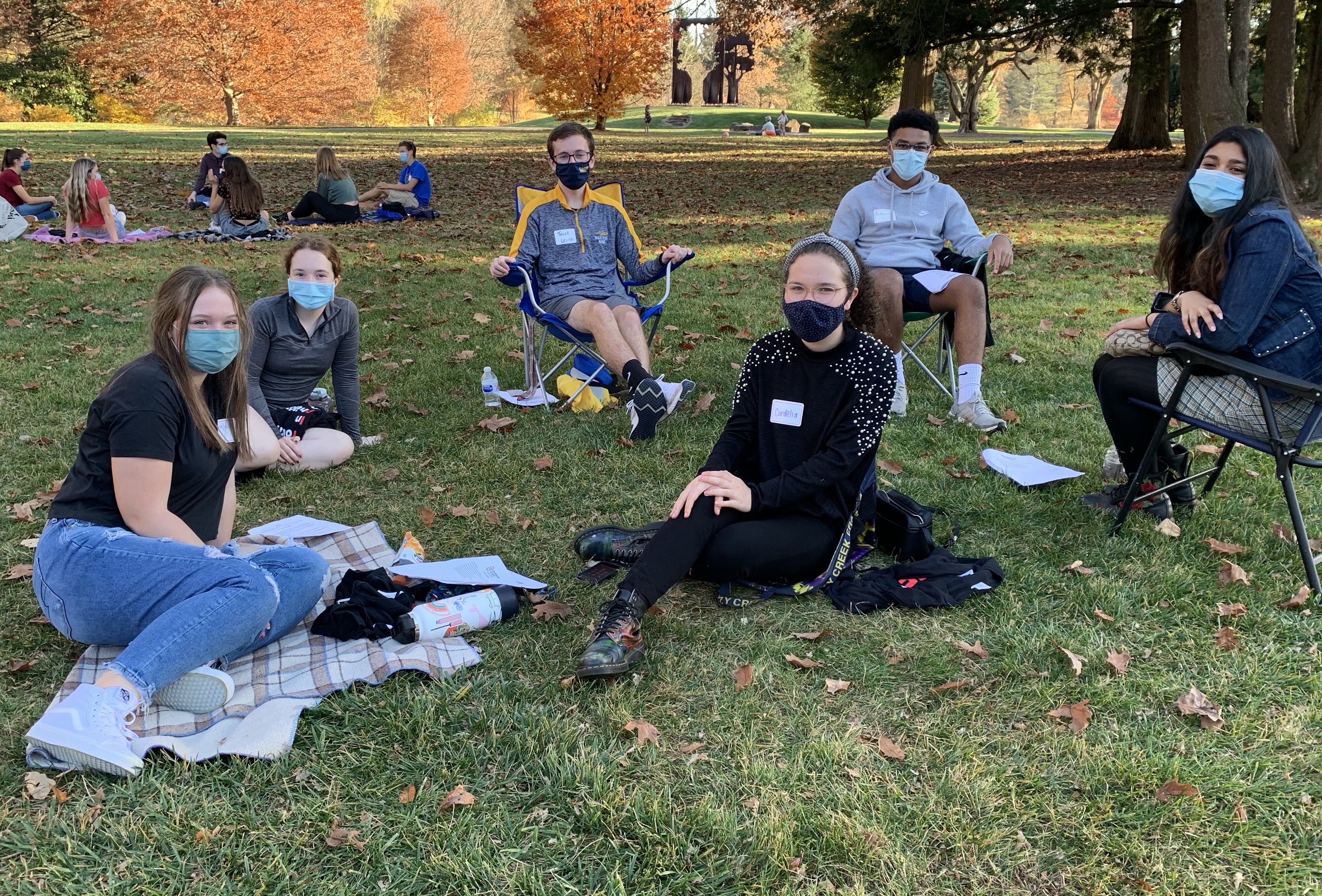 Students in face masks smile at the camera while sitting outside on an autumn day