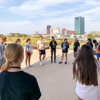 A group of students stand in a circle talking. They are outside and a cityscape stands behind them.